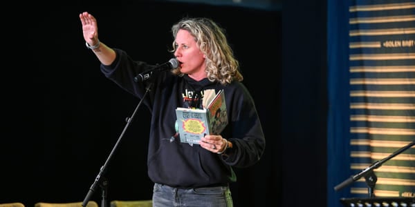 Kvinne med krøllete blondt hår som snakker i mikrofonen, holder en bok og rekker opp hånden. Iført svart genser og jeans.