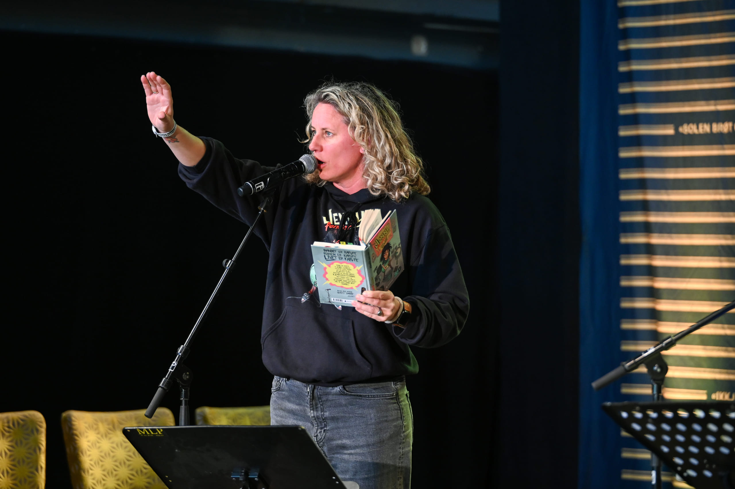 Kvinne med krøllete blondt hår som snakker i mikrofonen, holder en bok og rekker opp hånden. Iført svart genser og jeans.