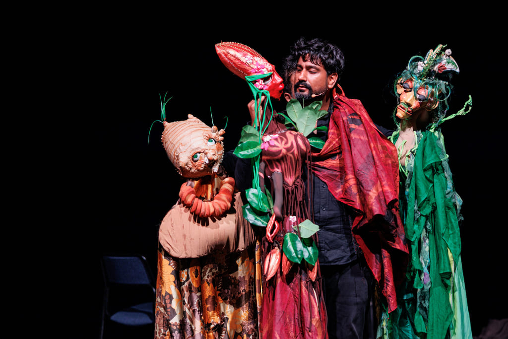 Utøver i rød kappe med tre fantastiske dukkevesener: plantelignende, løkhodet og skogånd, på mørk scene.