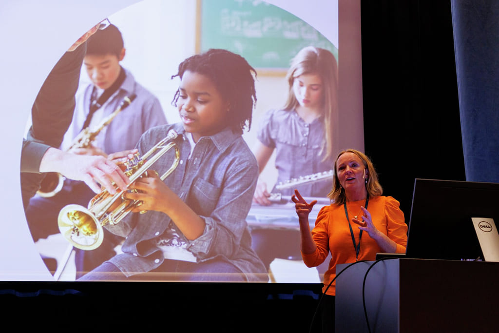 Presentatør i oransje skjorte gestikulerer ved siden av en projeksjon av elever som spiller musikkinstrumenter i et klasserom.