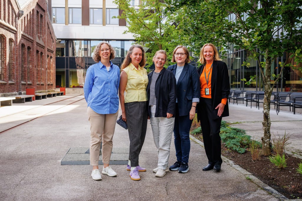 Fem smilende middelaldrende kvinner står sammen utenfor en bygning, kledd i uformelt forretningsantrekk med fargerike skjorter og jakker.