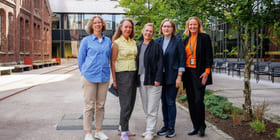 Fem smilende middelaldrende kvinner står sammen utenfor en bygning, kledd i uformelt forretningsantrekk med fargerike skjorter og jakker.