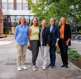 Fem smilende middelaldrende kvinner står sammen utenfor en bygning, kledd i uformelt forretningsantrekk med fargerike skjorter og jakker.