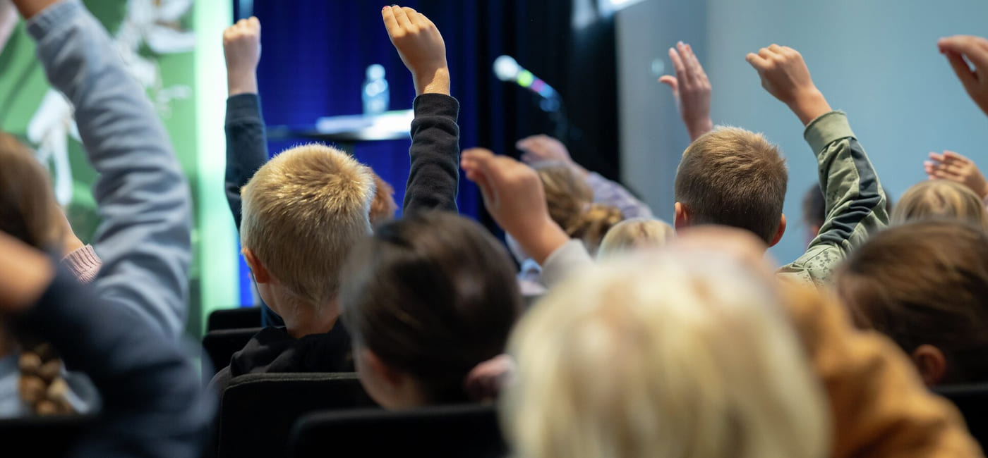 Elever med løftede hender i et publikum, vendt mot en scene med mikrofon og vannflaske synlig.