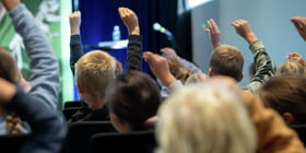 Elever med løftede hender i et publikum, vendt mot en scene med mikrofon og vannflaske synlig.