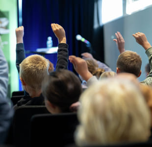 Elever med løftede hender i et publikum, vendt mot en scene med mikrofon og vannflaske synlig.