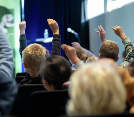 Elever med løftede hender i et publikum, vendt mot en scene med mikrofon og vannflaske synlig.