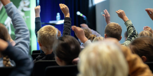 Elever med løftede hender i et publikum, vendt mot en scene med mikrofon og vannflaske synlig.