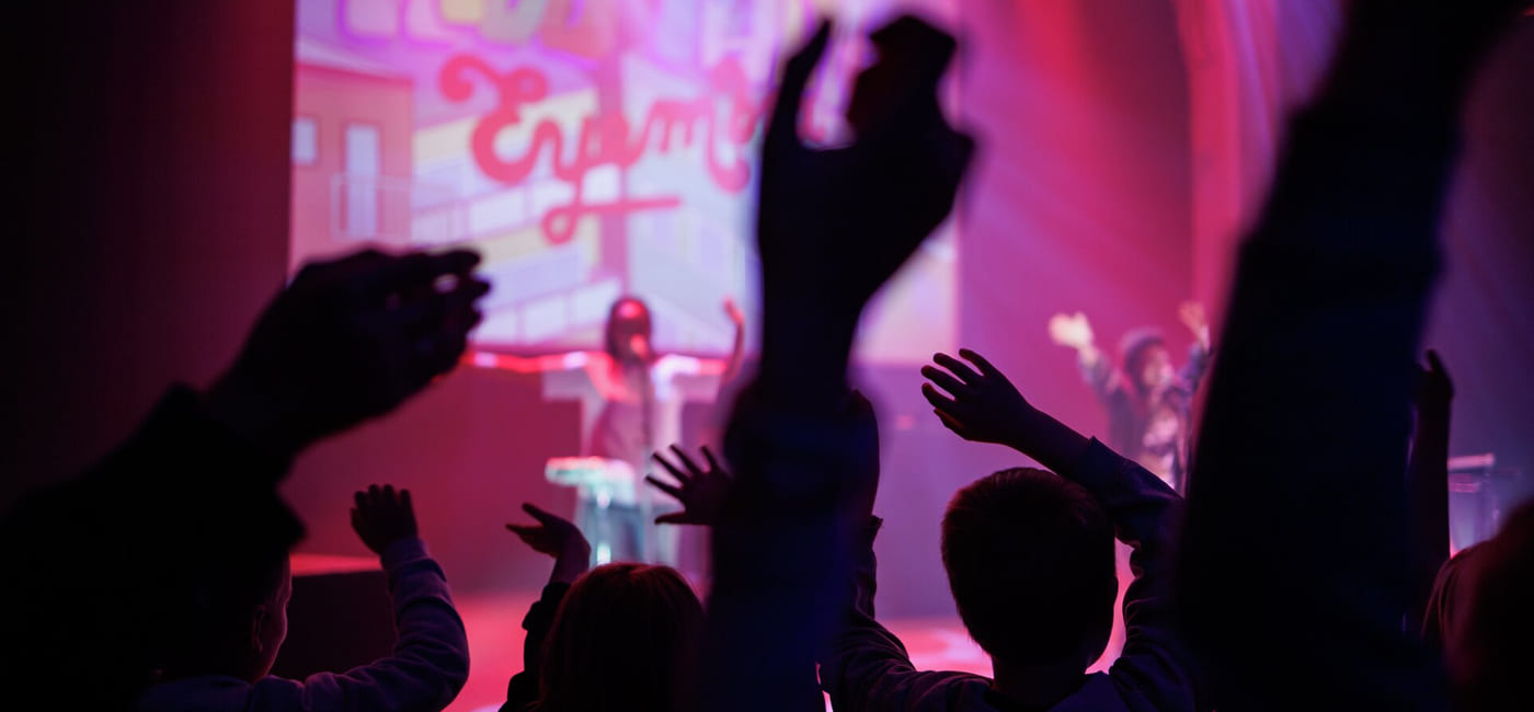 Konsertpublikum med hevede hender i silhuett mot rosa og lilla scenebelysning, utøvere synlig i bakgrunnen.