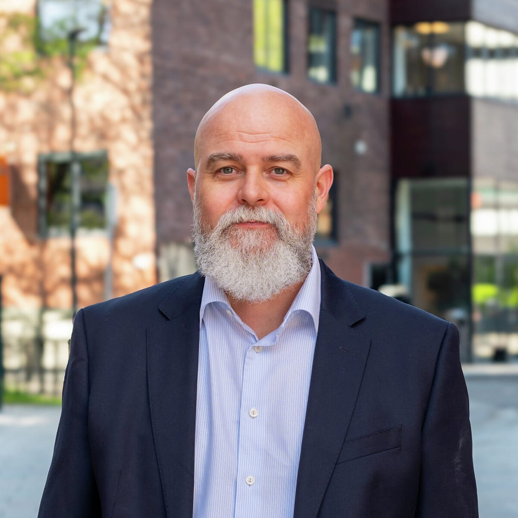 Skallet mann med hvitt skjegg iført mørk dress og lys skjorte står på fortauet foran murbygninger.