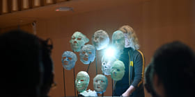 Kunstinstallasjon med opplyste, gjennomskinnelige masker på stativer, sett av tilskuere. En person i mørke klær står bak utstillingen.