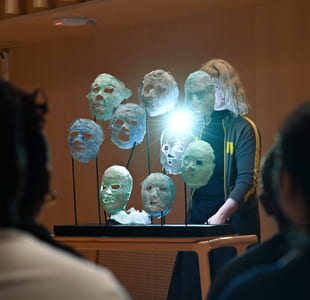 Kunstinstallasjon med opplyste, gjennomskinnelige masker på stativer, sett av tilskuere. En person i mørke klær står bak utstillingen.