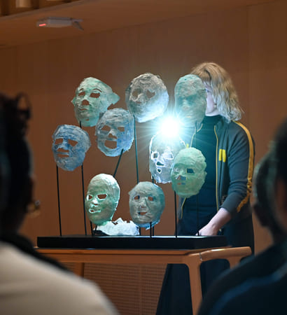 Kunstinstallasjon med opplyste, gjennomskinnelige masker på stativer, sett av tilskuere. En person i mørke klær står bak utstillingen.