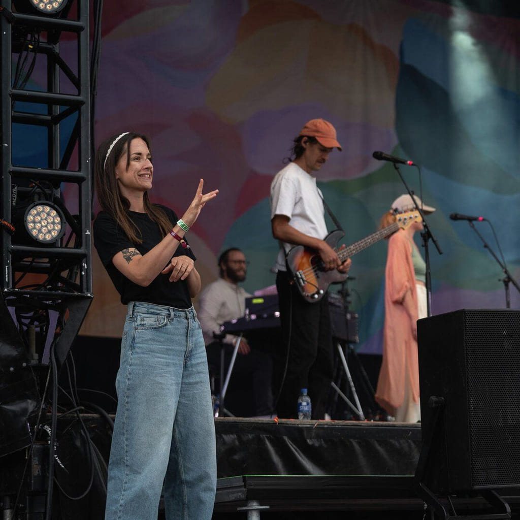 Et band står på en scene. En kvinne i hvit kjole og rosa caps synger. Til venstre står en kvinne og tegnspråktolker.