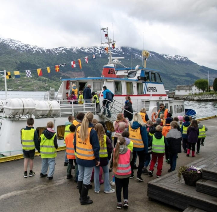 En mellomstor båt pyntet med fargerike flagg ligger til kai i et fjordlandskap. Flere barn og lærere i refleksvester er på vei ombord i båten.