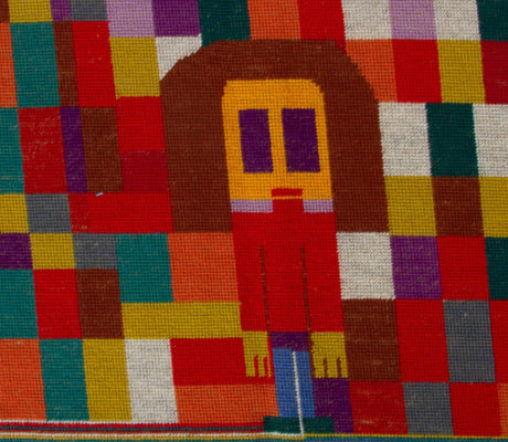 Et fargerikt vevd teppe. I midten ses en menneskefigur.