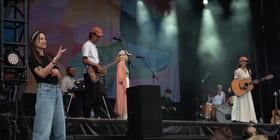 Et band står på en scene. En kvinne i hvit kjole og rosa caps synger. Til venstre står en kvinne og tegnspråktolker.