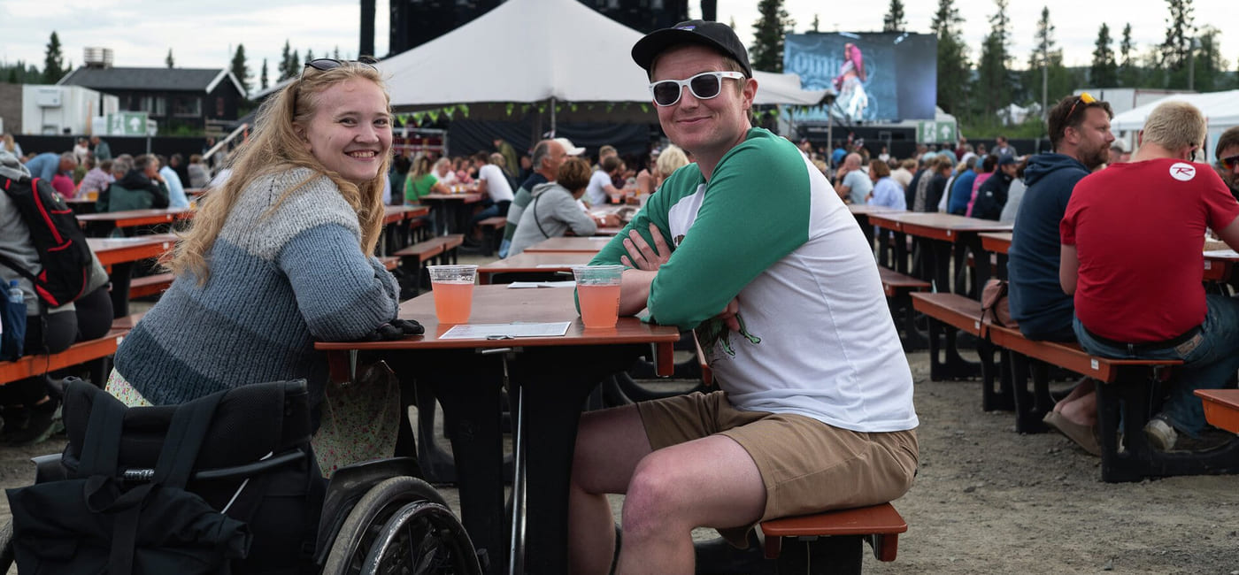 Et par sitter ved en benk på et festivalområde. De smiler i kamera. Hun sitter i rullestol.