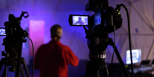 Videokameraer på stativ som filmer en person i rødt antrekk mot et lilla opplyst bakteppe i et produksjonsstudio.