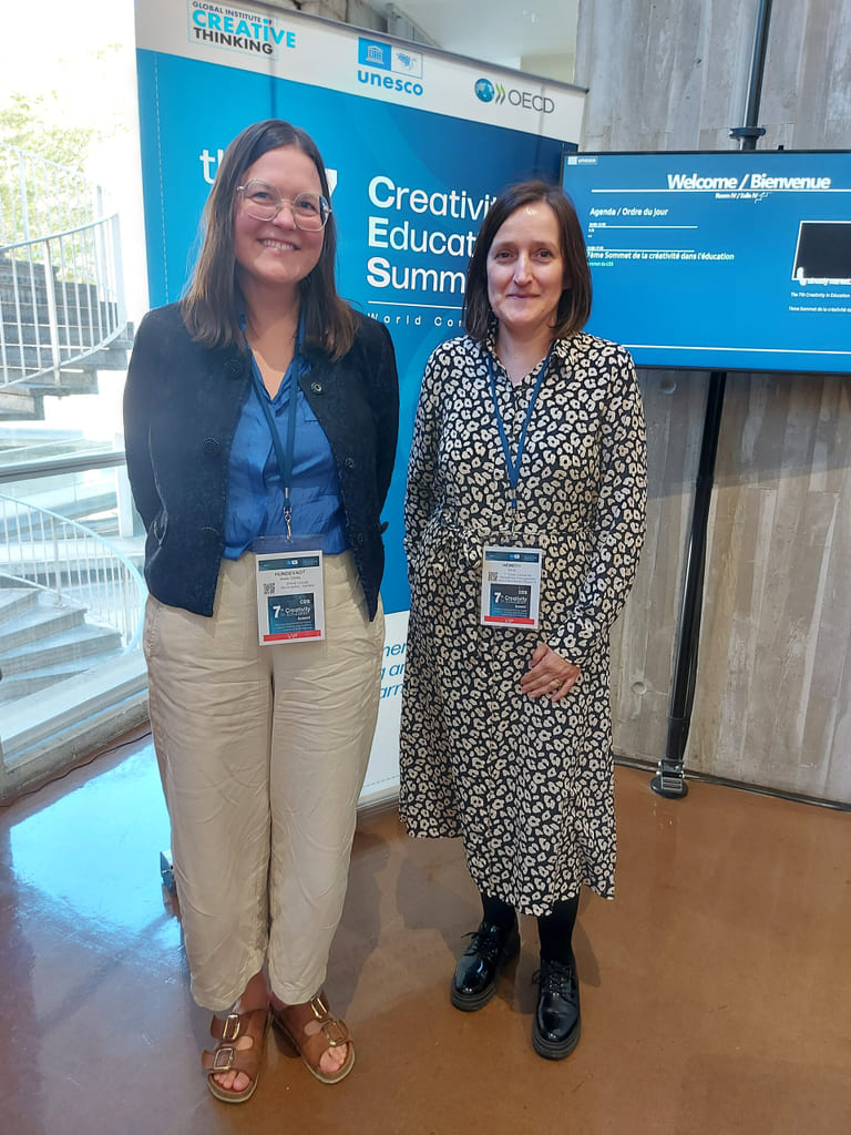 To smilende kvinner på Creativity in Education Summit, står foran bannere for arrangementet. Den ene har på seg blått, den andre i mønstret kjole.