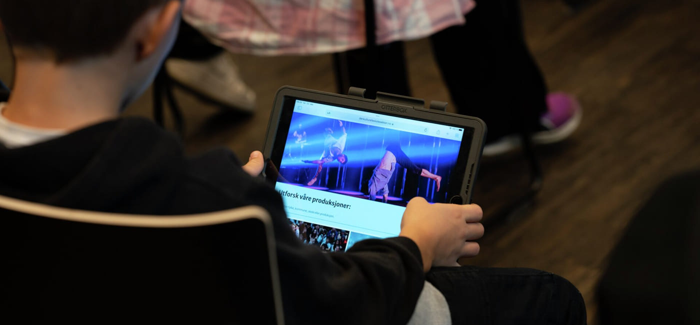 Barn som bruker nettbrett som viser nettside for Den kulturelle skolesekken, sittende i et svakt opplyst rom med en annen person i nærheten.