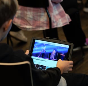 Barn som bruker nettbrett som viser nettside for Den kulturelle skolesekken, sittende i et svakt opplyst rom med en annen person i nærheten.