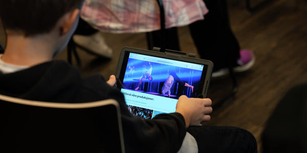 Barn som bruker nettbrett som viser nettside for Den kulturelle skolesekken, sittende i et svakt opplyst rom med en annen person i nærheten.