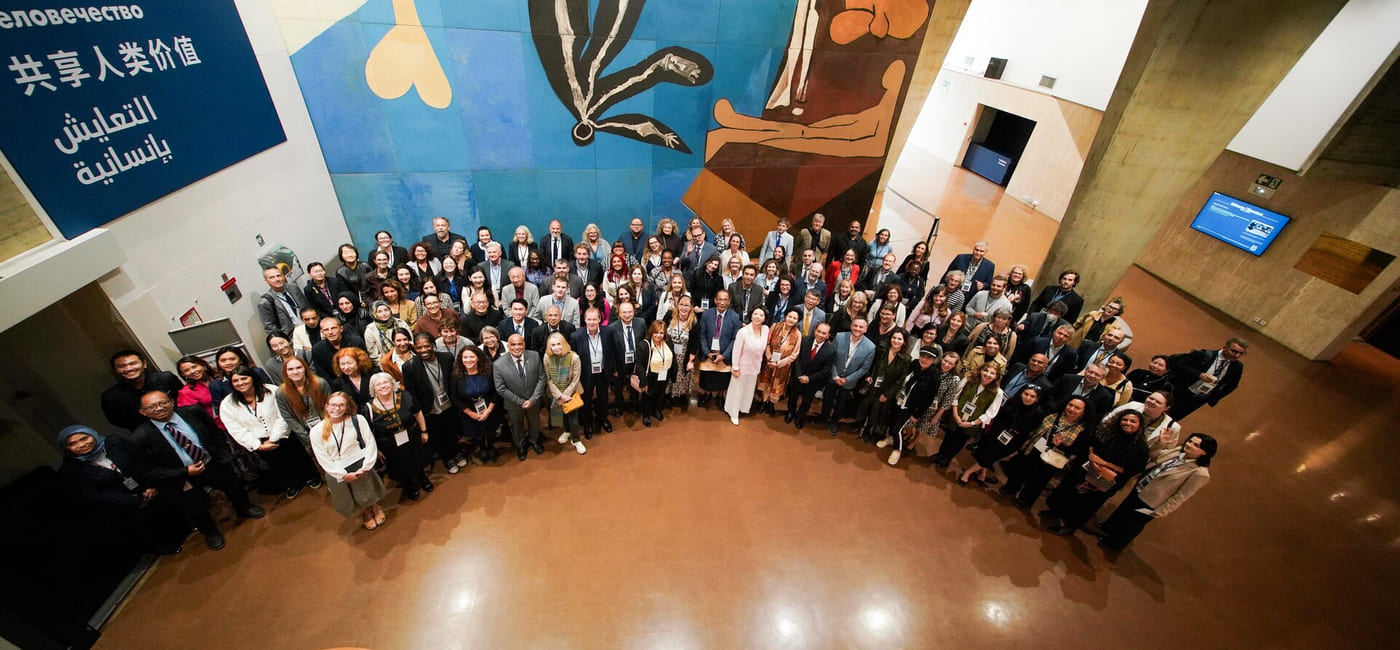 Stort gruppebilde av ulike deltakere på en internasjonal konferanse, med fargerikt abstrakt veggmaleri og flerspråklig skilt i bakgrunnen.