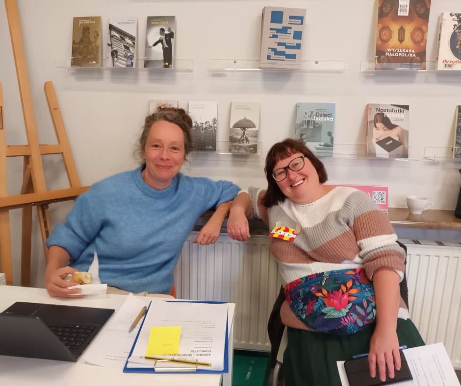 o personer sitter ved bord med tegne- og maleutstyr, omgitt av kunstverk på veggen