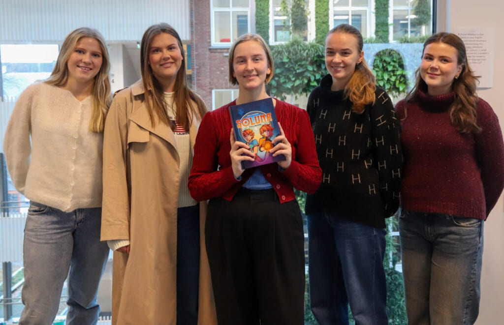 Fem personer står sammen innendørs, én holder en fargerik bok.