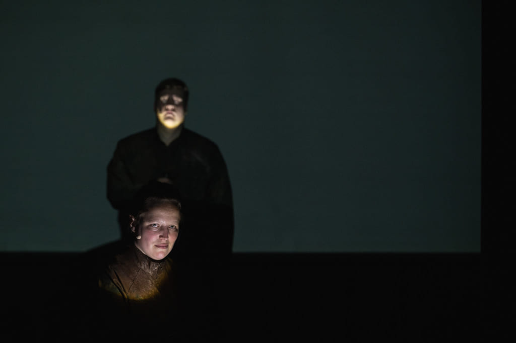 Person på mørk scene med spotlight. Bak henne skimtes en annen person.