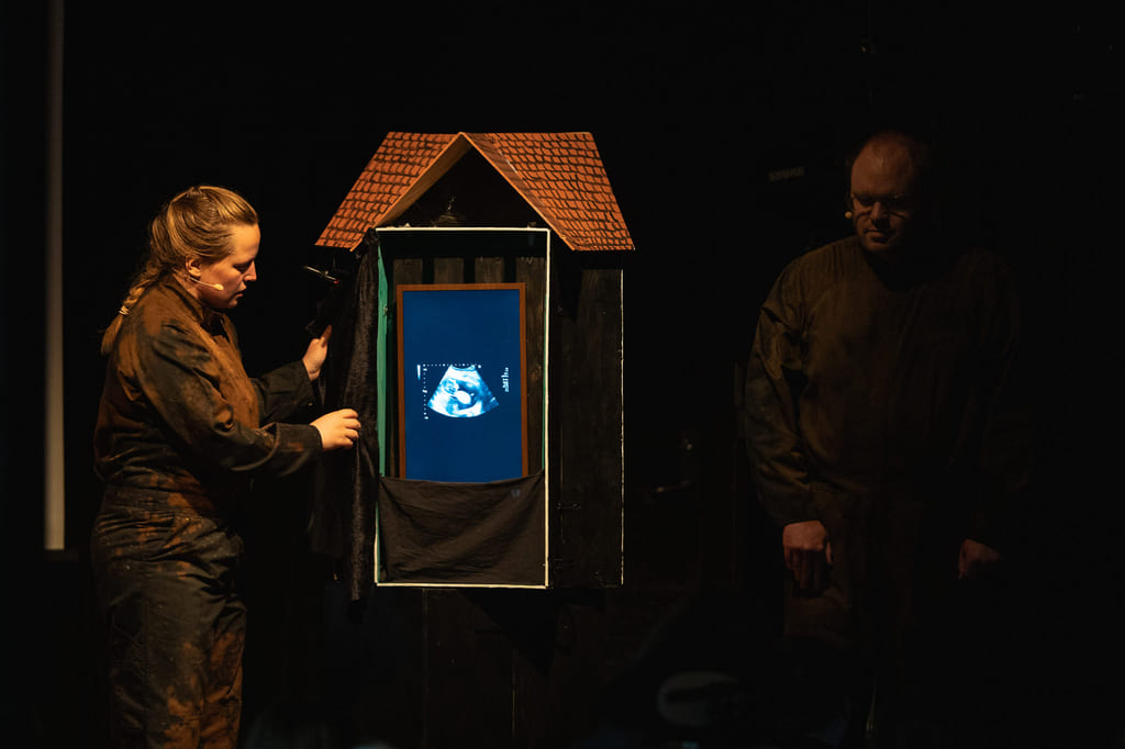 To personer på en mørk scene. I et dukkehus skimtes et ultralyd-bilde