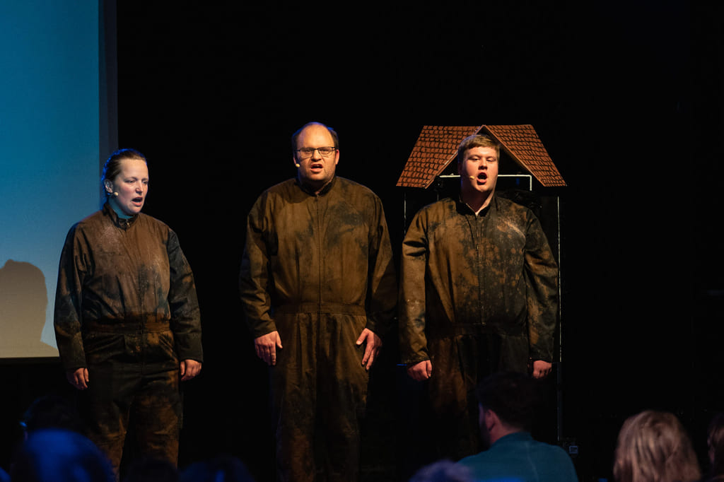 Flere personer på scene med mørk bakgrunn, iført kostymer