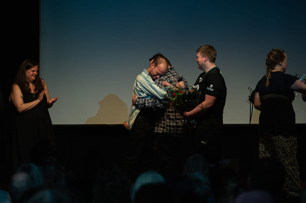 Fem personer på mørk scene, en holder den andre i armene