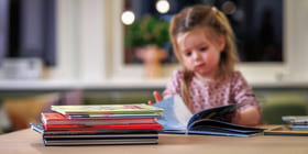 Et barn sitter og leser ved et bord. I forgrunnen er en bunke barnebøker.