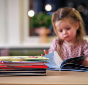 Et barn sitter og leser ved et bord. I forgrunnen er en bunke barnebøker.