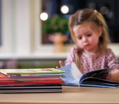 Et barn sitter og leser ved et bord. I forgrunnen er en bunke barnebøker.