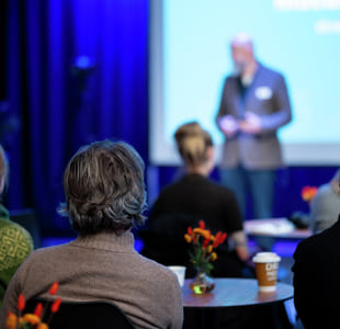 Konferansedeltakere sitter ved bord vendt mot en programleder på scenen med projeksjonsskjerm i et svakt opplyst blått rom.