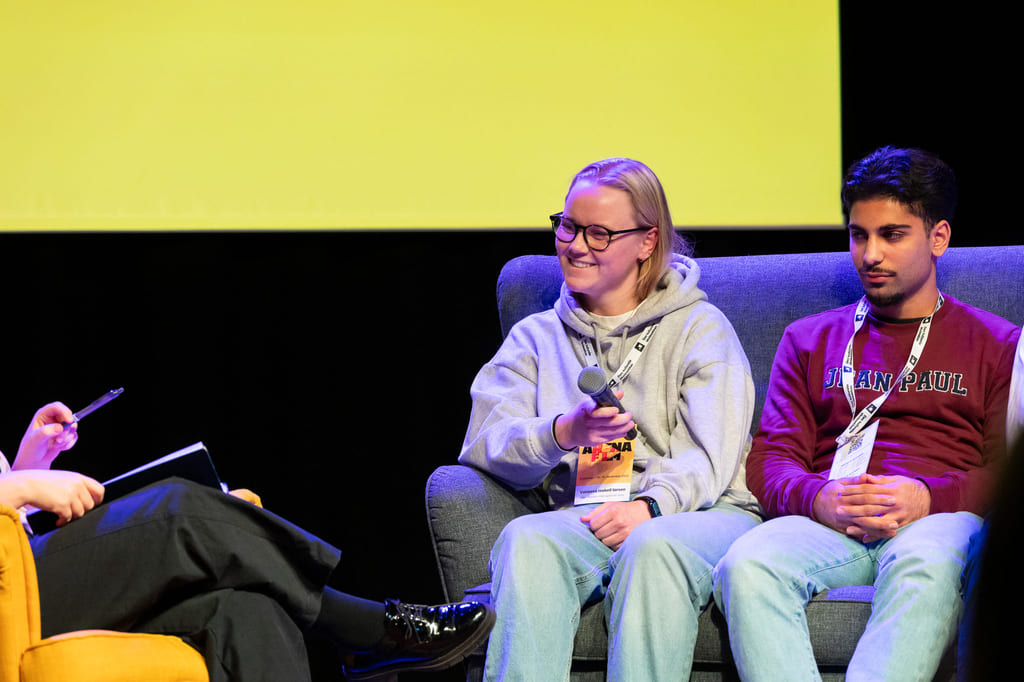 Paneldiskusjon med tre deltakere: intervjuer, smilende kvinne i hettegenser som holder mikrofon, og mann i DePaul-genser på scenen.