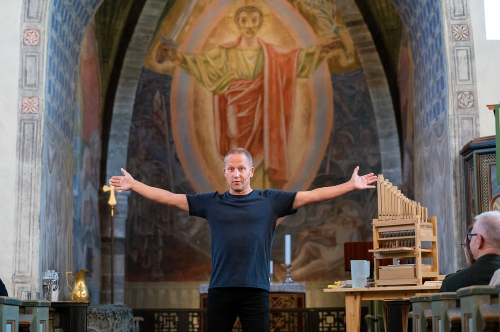 Mann med utstrakte armer står foran et stort religiøst veggmaleri i kirken, med et lite orgel synlig ved siden av ham.