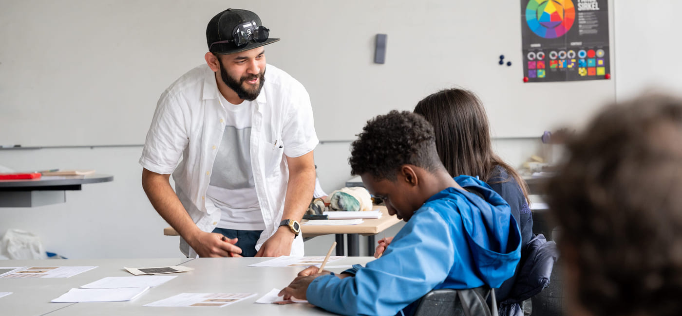 Person i hvit skjorte og lue hjelper to elever ved et pult i et klasserom med en fargerik plakat.
