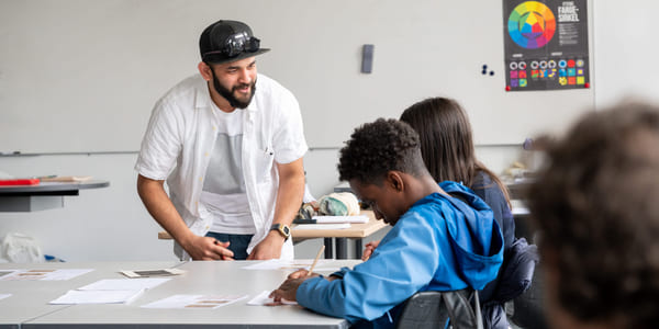 Person i hvit skjorte og lue hjelper to elever ved et pult i et klasserom med en fargerik plakat.