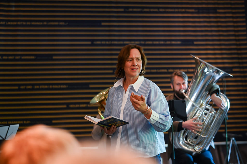 Kvinne holder en bok og gestikulerer. Mann i bakgrunnen spiller tuba. Stripete vegg bak dem. Musikalsk opptreden.