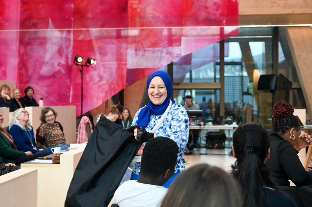 Kvinne i blå hijab og blomstrete topp snakker til et mangfoldig publikum i et fargerikt arrangementslokale med rosa bakgrunn.