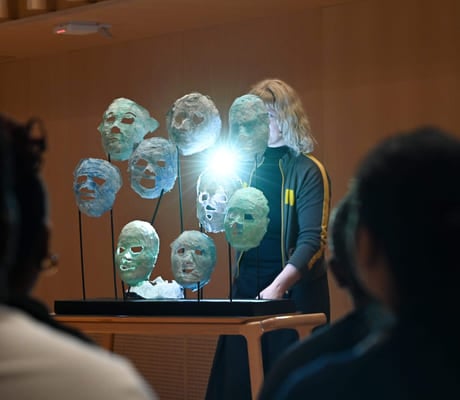 Kunstinstallasjon med opplyste, gjennomskinnelige masker på stativer, sett av tilskuere. En person i mørke klær står bak utstillingen.