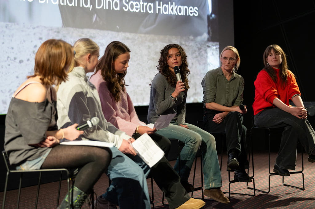Paneldiskusjon med seks kvinner sittende på scenen. Én snakker inn i mikrofonen.