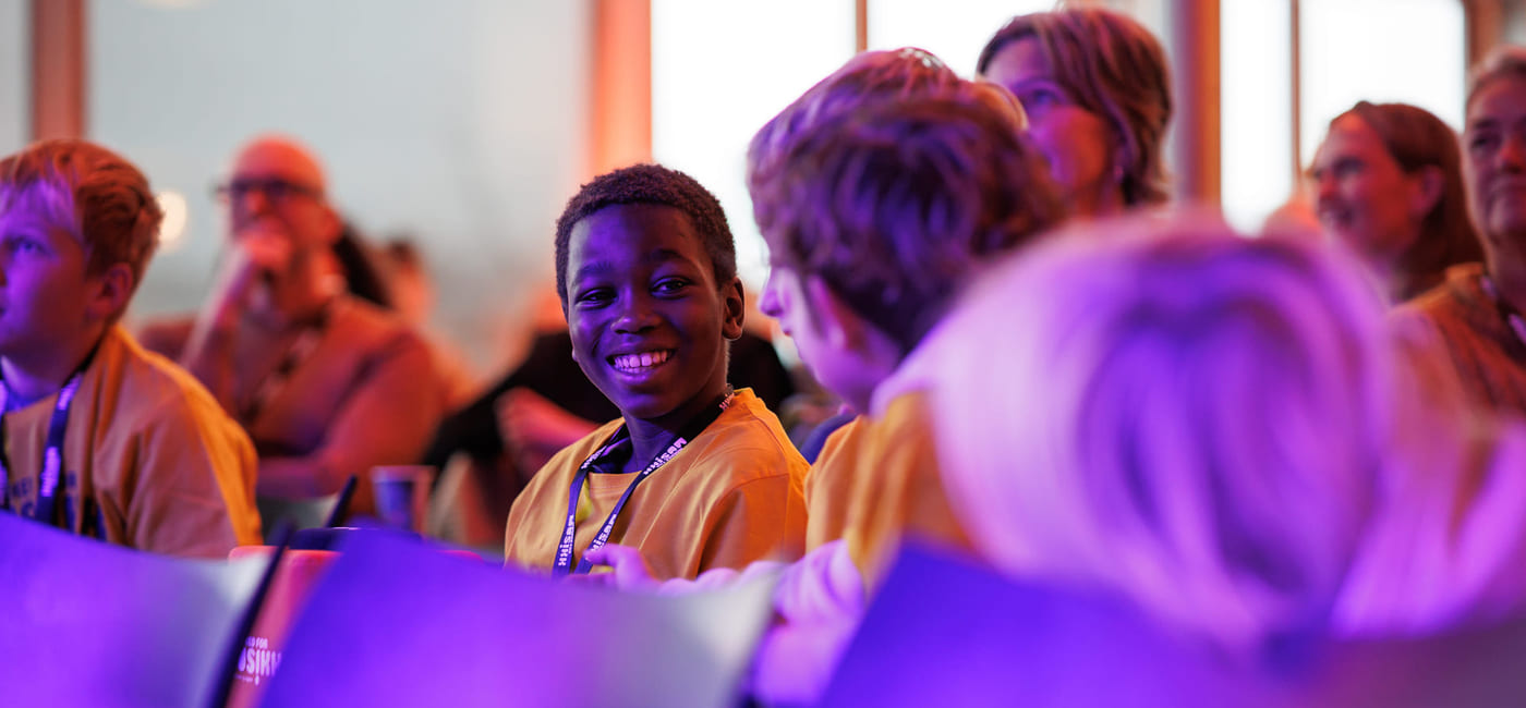 Gruppe mennesker på arrangementet, ung person i gul skjorte som smiler sterkt, badet i lilla lys.