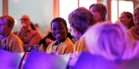 Gruppe mennesker på arrangementet, ung person i gul skjorte som smiler sterkt, badet i lilla lys.