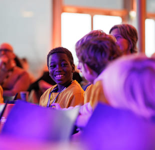 Gruppe mennesker på arrangementet, ung person i gul skjorte som smiler sterkt, badet i lilla lys.