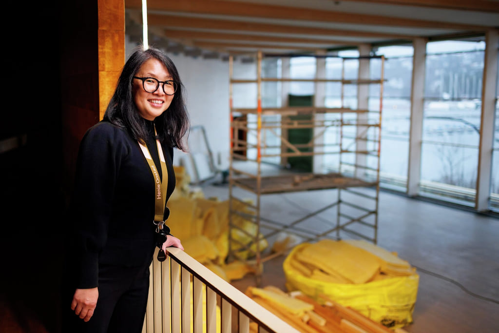 Forretningskvinne i svart blazer smiler mens hun står ved en moderne kontorresepsjonsdisk med gule detaljer.